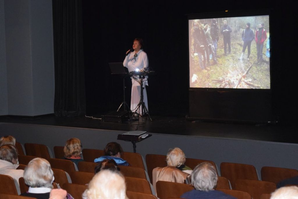 Žmonės, sėdintys salėje. Lektorė su mikrofonu.