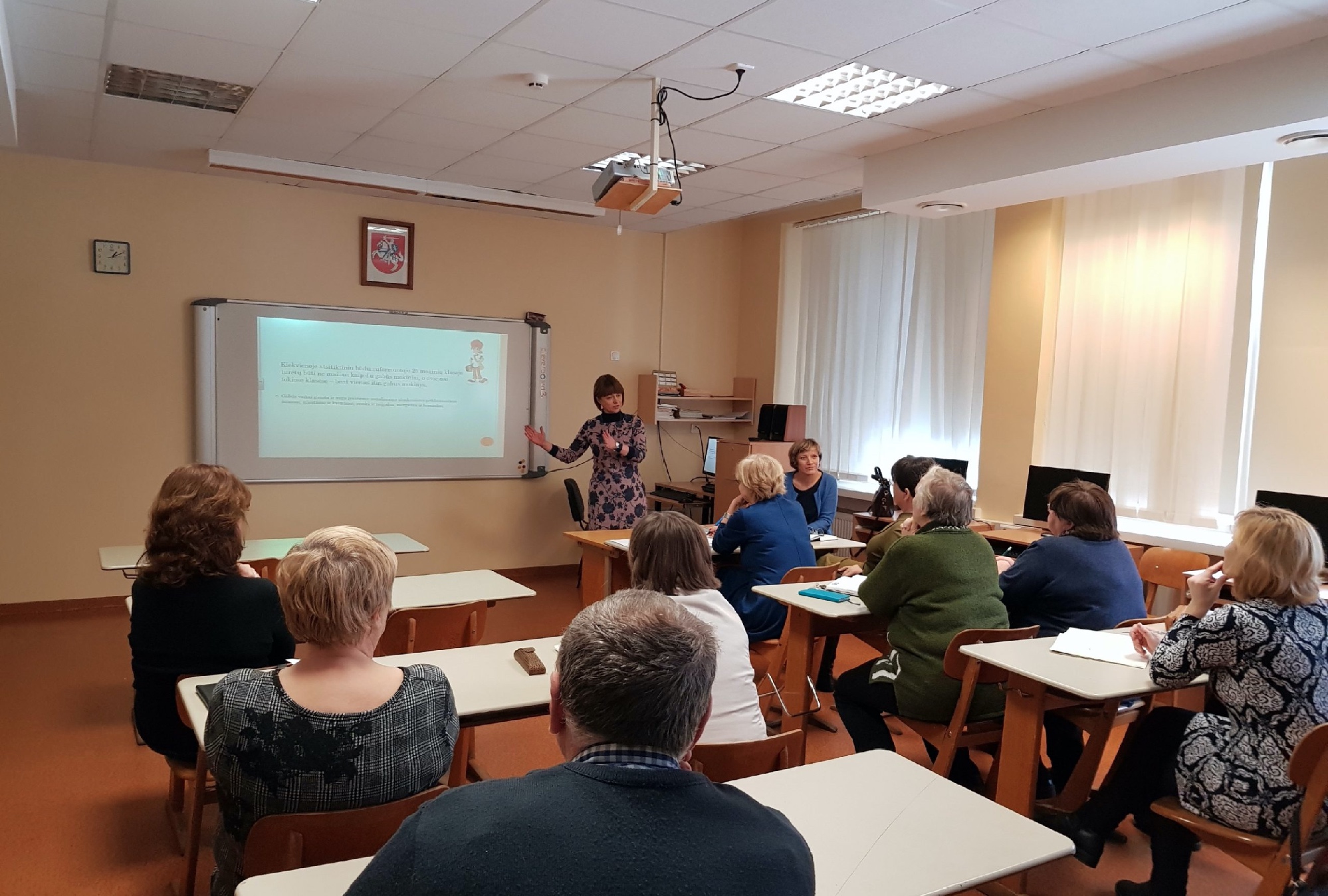 Mokymų dalyviai, susėdę klasėje, stebi lektorės demonstruojamą skaidrę ekrane