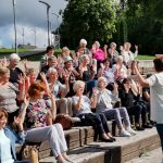 Ant laiptelių susėdę žmonės, iškėlę rankas sportuoja, žiūrėdami į trenerę