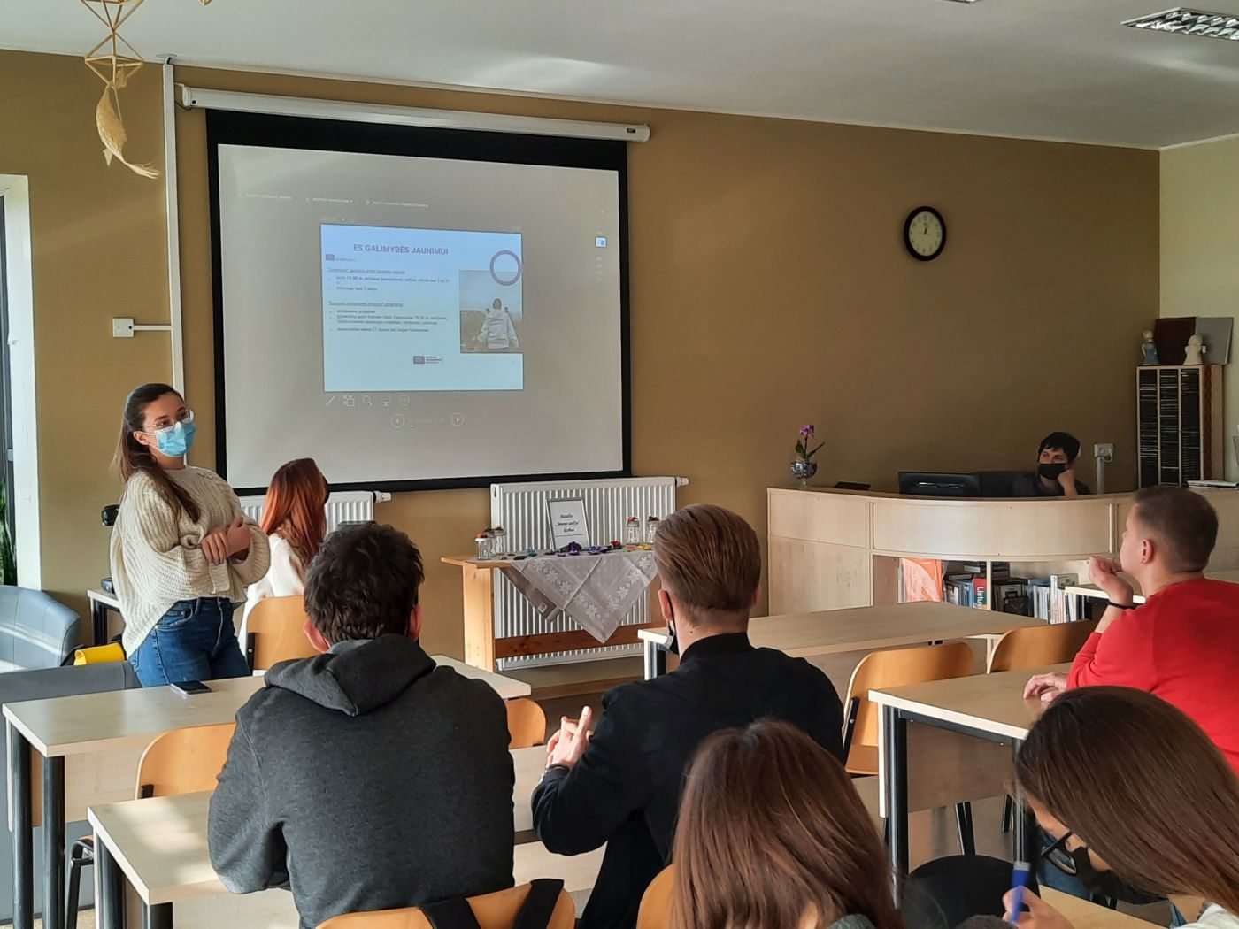 Jauni žmonės, sėdintys klasėje. Lektorė stovi atsisukusi į auditoriją