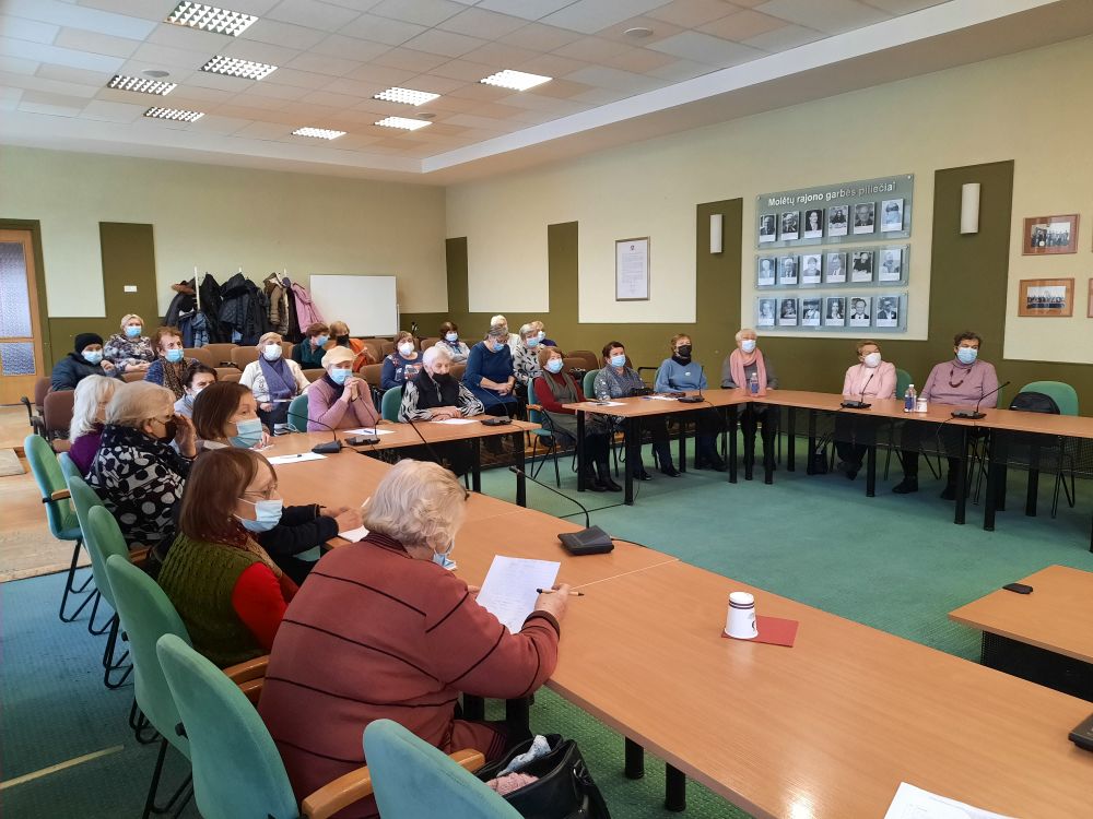 Prie stalų ratu sėdintys žmonės