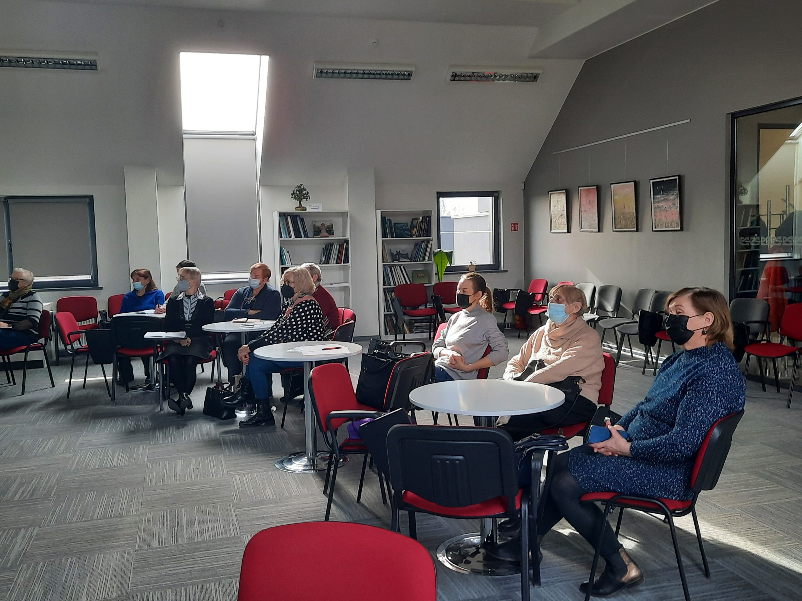 Salėje prie staliukų susėdę žmonės klausosi paskaitos