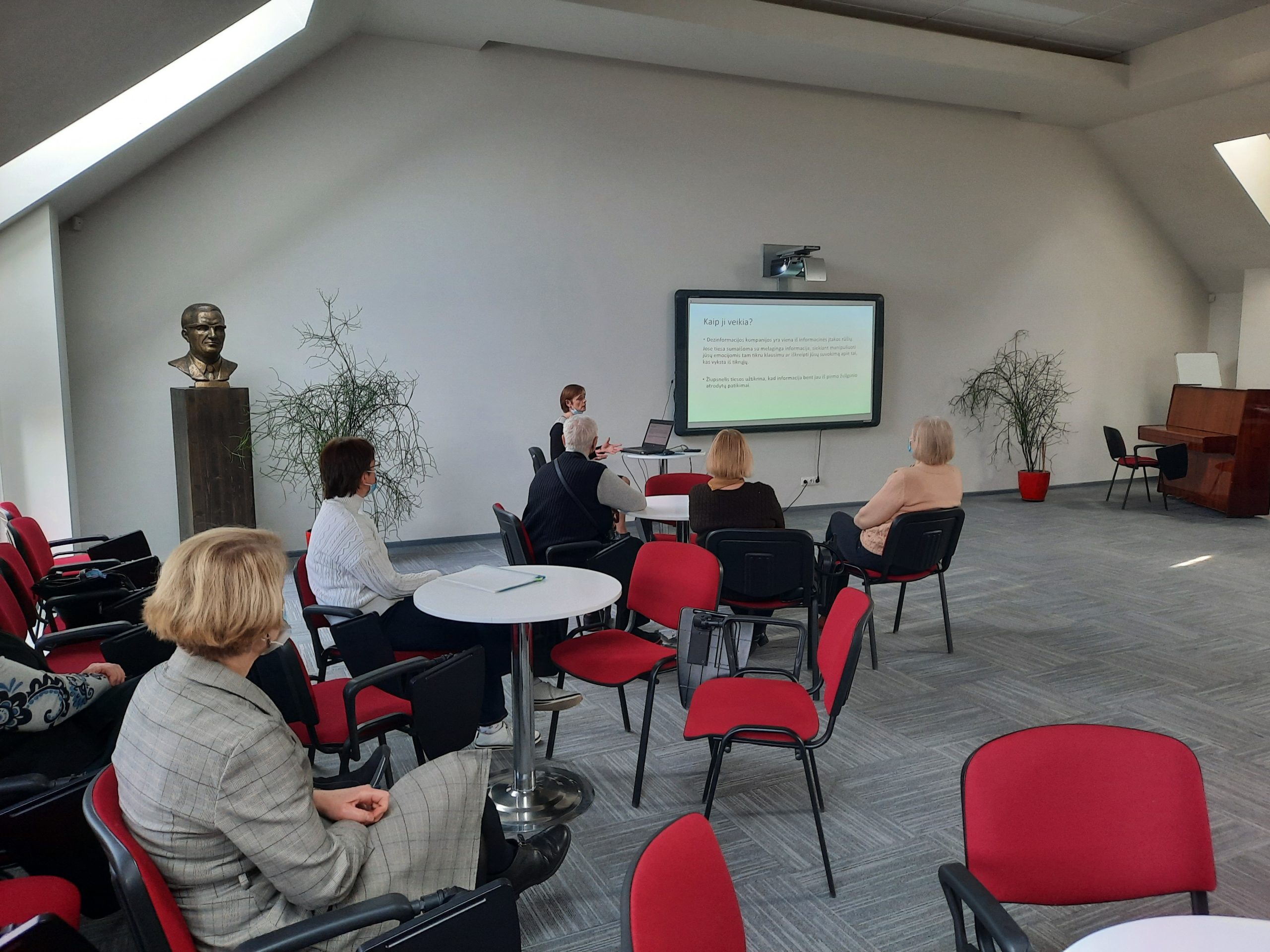 Salėje prie staliukų susėdę žmonės klausosi paskaitos