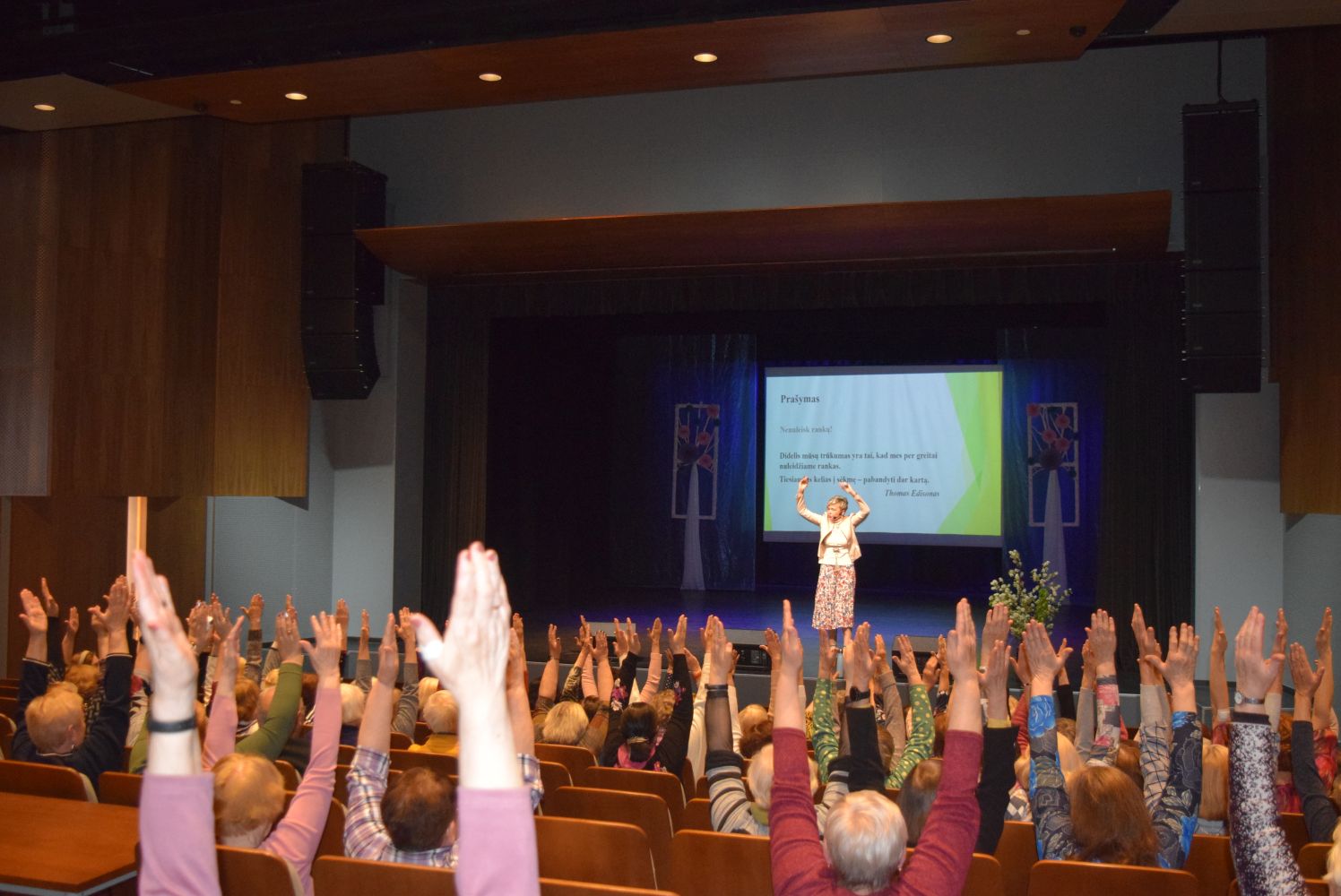 Salėje sėdintys paskaitos dalyviai, visi iškėlę abi rankas į viršų